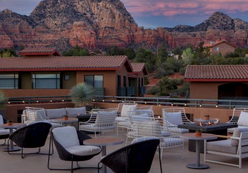 A rooftop patio with seating and fire pits, surrounded by buildings and stunning red rock mountains in the background, under a pinkish sky.