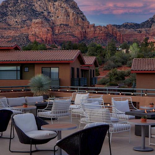 A rooftop patio with seating and fire pits, surrounded by buildings and stunning red rock mountains in the background, under a pinkish sky.