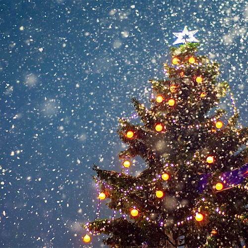 A decorated Christmas tree with lights and a star topper stands under falling snowflakes against a clear blue sky.