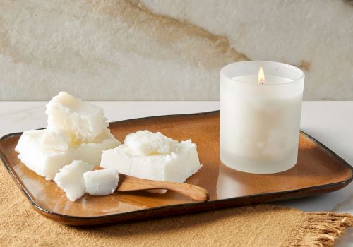 A lit candle in a frosted glass sits on a tray with chunks of wax and a wooden spoon on a cloth.