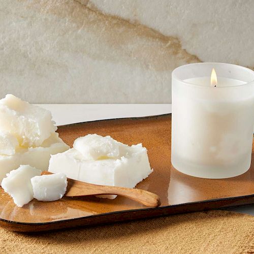 A lit candle in a frosted glass sits on a tray with chunks of wax and a wooden spoon on a cloth.