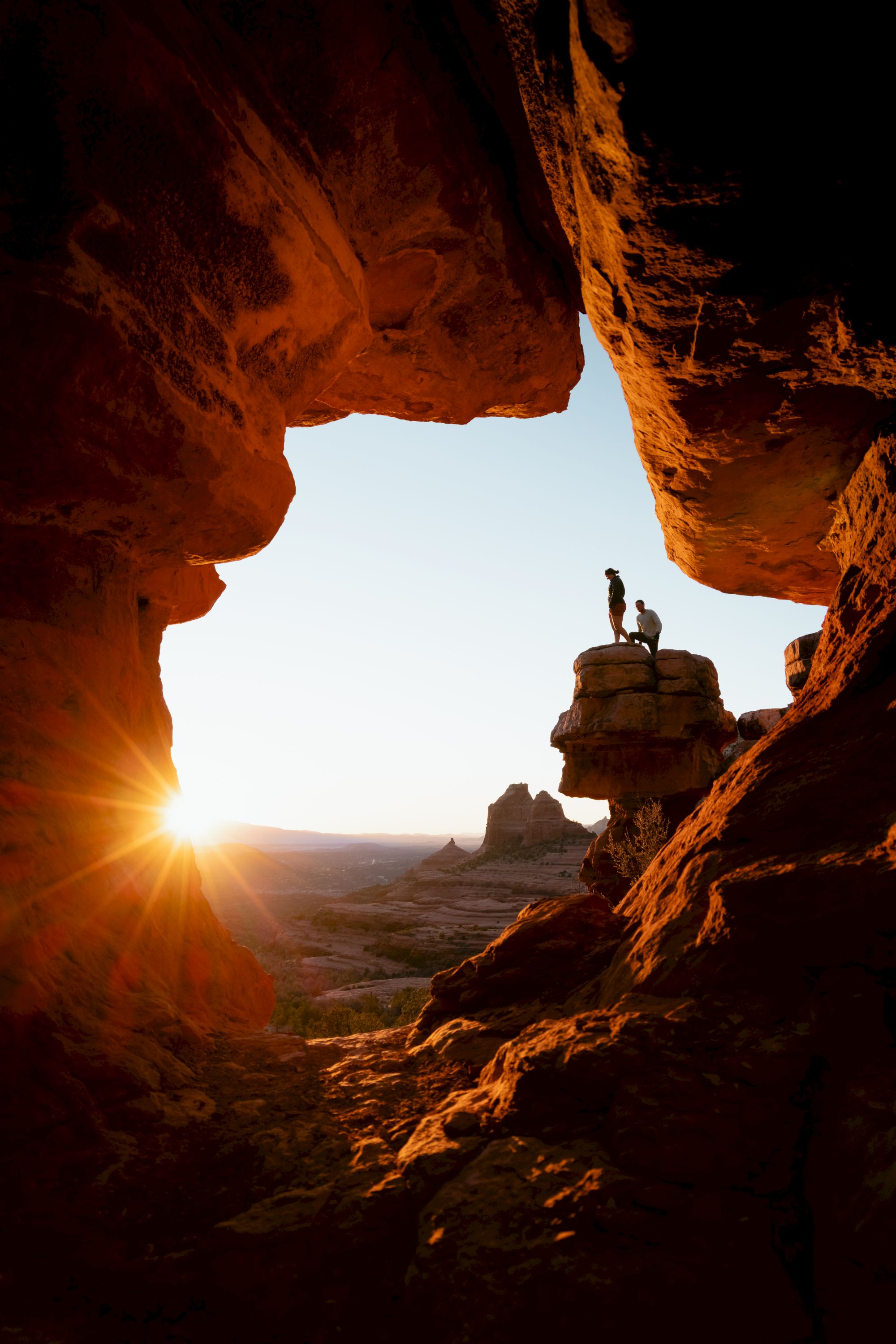 A sunlit canyon with rock formations, two people stand on an outcrop silhouetted against the sky, capturing an adventurous moment.