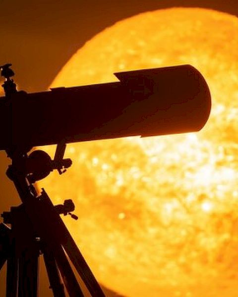 A telescope is silhouetted against a large, bright image of the sun in a dramatic sunset scene.